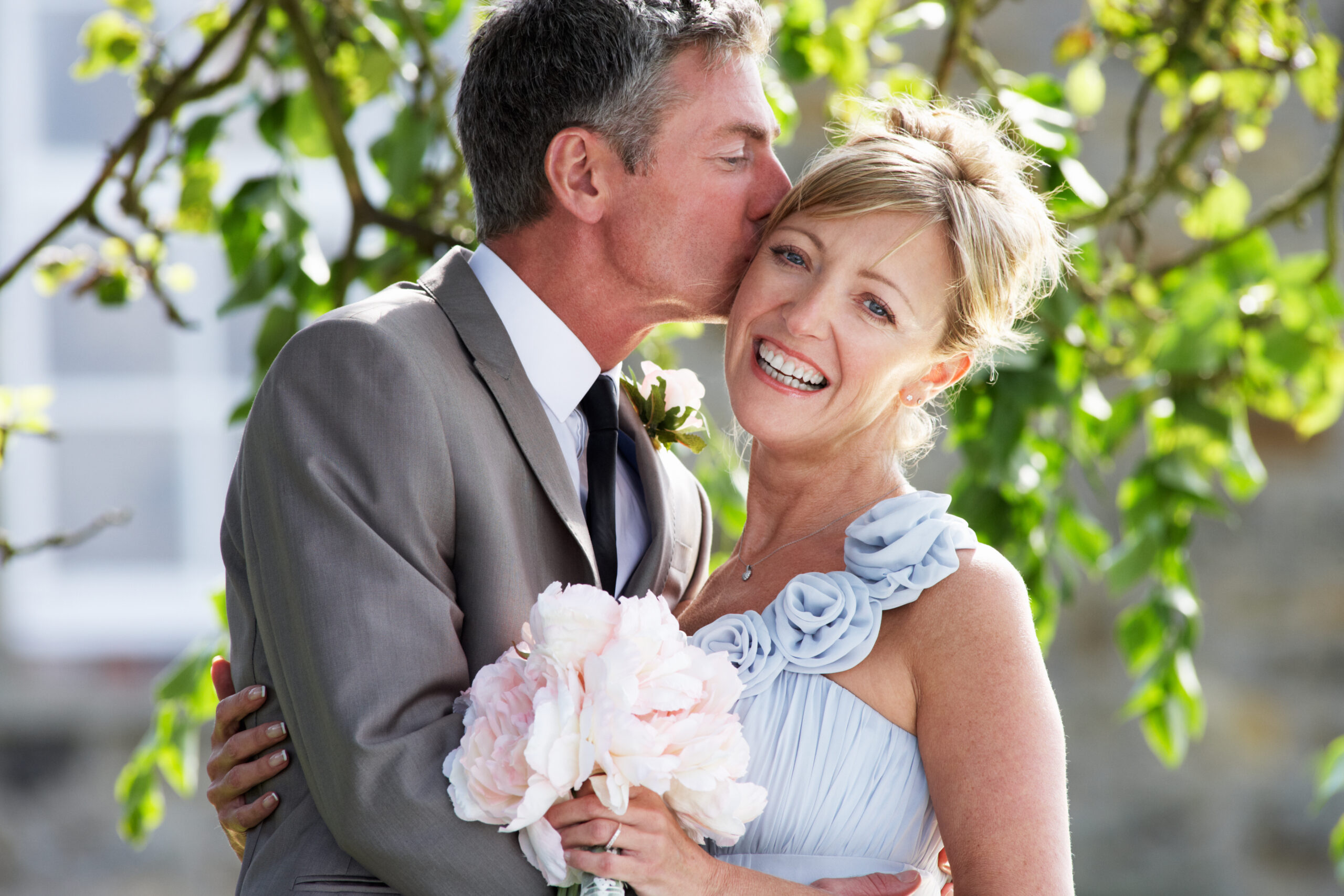 Romantic Bride And Groom Embracing Outdoors Kissing And Smiling To Camera